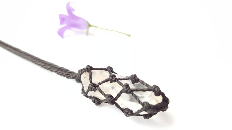 May include: A clear quartz crystal is wrapped in a black macrame cord, creating a pendant necklace. The crystal is long and slender, with a pointed tip. The macrame cord is tied in a intricate pattern, creating a delicate and elegant design.