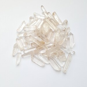 May include: A pile of clear quartz crystal points, also known as rock crystals, on a white background. The crystals are various sizes and shapes, with some having a slight golden hue.