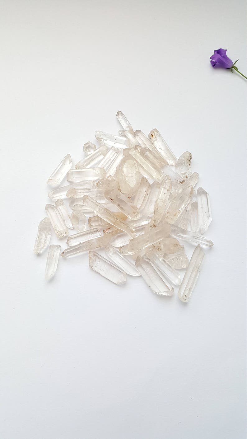 May include: A pile of clear quartz crystal points, also known as rock crystals, on a white background. The crystals are various sizes and shapes, with some having a slight golden hue.