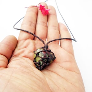 Può includere: Una collana fatta a mano con un ciondolo in pietra verde e rossa in una rete intrecciata marrone, sospeso a un cordino marrone. Un piccolo fiore rosso è posto vicino alla collana.