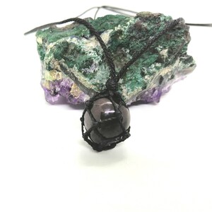 Può includere: Collana con ciondolo in pietra nera, cordoncino nero e gabbia in macramé nero. Il ciondolo è su una roccia viola e verde.