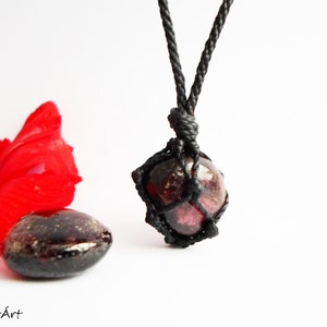 May include: A dark red gemstone pendant necklace with a black cord. The gemstone is wrapped in a black cord macrame knot.