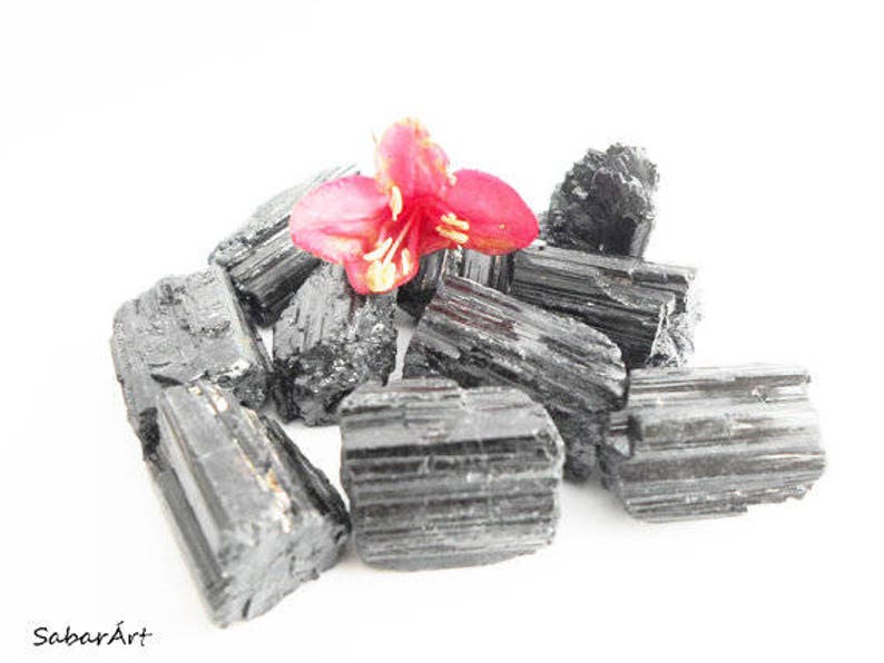 May include: A pile of black tourmaline crystals with a single pink flower on top. The crystals are rough and have a natural, unpolished look.
