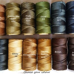 Op de afbeelding: Twaalf spoelen gewaxt garen in verschillende kleuren, waaronder bruin, groen, blauw, zwart en goud. Elke spoel heeft een label met de kleurnaam en een nummer.