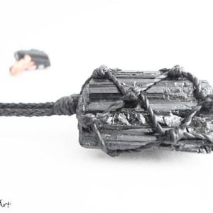 May include: Black Tourmaline crystal wrapped in black cord. The crystal is a dark grey color and has a rough, natural texture. The cord is tied in a knot at the top of the crystal.