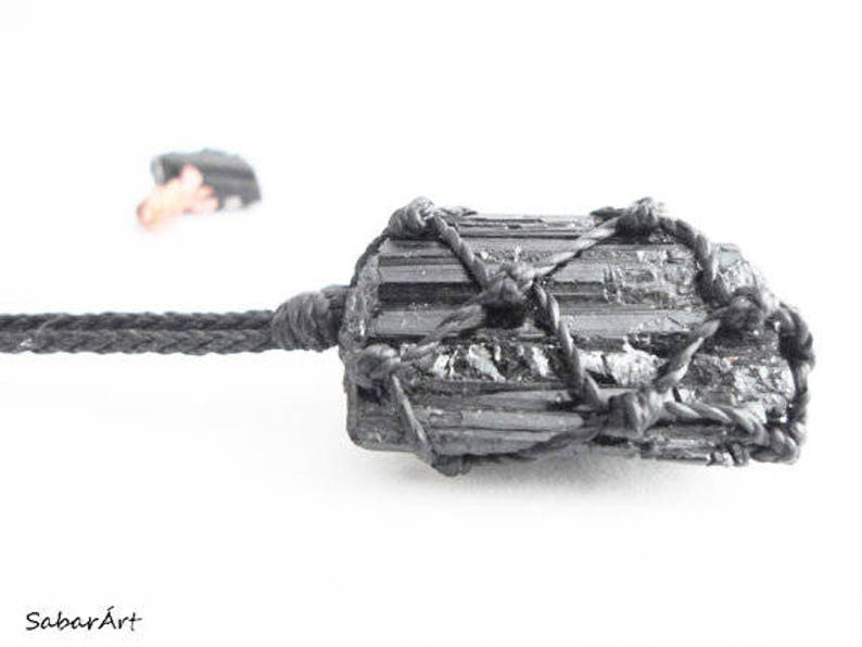 May include: Black Tourmaline crystal wrapped in black cord. The crystal is a dark grey color and has a rough, natural texture. The cord is tied in a knot at the top of the crystal.