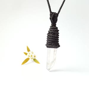May include: A clear quartz crystal pendant with a black macrame knot. The pendant is hanging from a black cord.