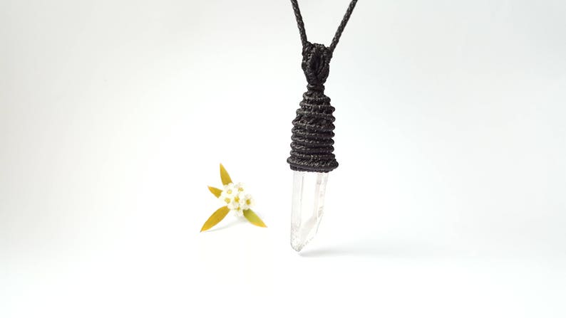 May include: A clear quartz crystal pendant with a black macrame knot. The pendant is hanging from a black cord.