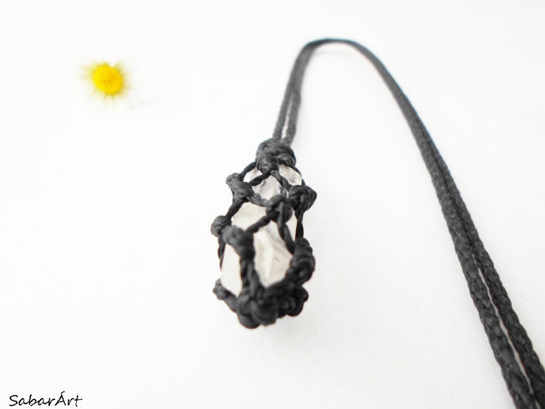 May include: A white crystal is encased in a black macrame net, hanging from a black cord. The crystal is a natural, irregular shape.