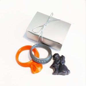May include: A silver gift box tied with a silver ribbon, with two orange and gray rings and a chocolate figurine of a couple.