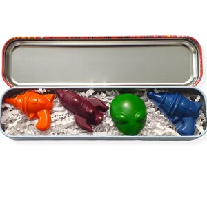 May include: A set of four space-themed crayons in a tin box. The crayons are shaped like a rocket, an alien, a gun, and a spaceship. The crayons are red, green, blue, and orange.