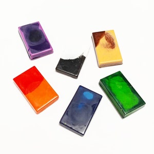 May include: Six rectangular blocks of wax crayons in various colors, including purple, black, orange, blue, green, and yellow. The crayons are arranged on a white surface.