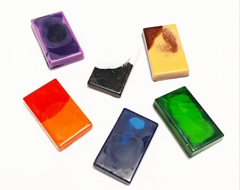 May include: Six rectangular blocks of wax crayons in various colors, including purple, black, orange, blue, green, and yellow. The crayons are arranged on a white surface.