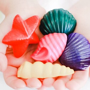 May include: A child's hand holds a collection of colorful seashell-shaped crayons. The crayons are red, green, pink, purple, and yellow.