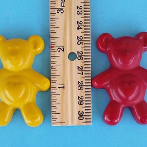 May include: Two small teddy bear shaped crayons, one yellow and one red, with a heart shape on their chest. The crayons are next to a ruler measuring 3 inches.