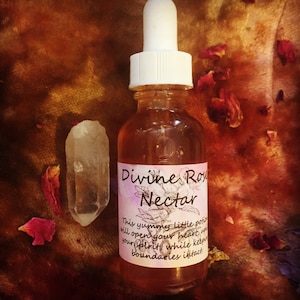Könnte beinhalten: Eine klare Glasflasche mit weißem Tropfer, beschriftet mit "Divine Rose Nectar". Die Flasche enthält eine hellbernsteinfarbene Flüssigkeit. Ein klarer Quarzkristall und Rosenblätter liegen neben der Flasche.
