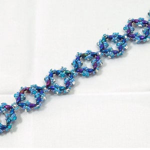 May include: A beaded bracelet featuring square-shaped links. The bracelet is adorned with blue and purple beads, creating a shimmering effect. The clasp is silver-toned. The bracelet is displayed on a white surface.