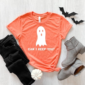 May include: A coral orange t-shirt with a white ghost graphic and the text "Can I keep you?" printed on the front. The shirt is lying on a white wooden surface with black jeans, grey ankle boots, and a white pumpkin.