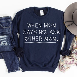 Könnte beinhalten: Marineblaues Sweatshirt mit dem weißen Text "WHEN MOM SAYS NO, ASK OTHER MOM". Das Bild zeigt auch blaue Jeans, einen grauen Hut und einen floralen Schal.