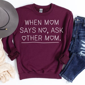 Könnte beinhalten: Ein weinrotes Sweatshirt mit dem weißen Text "WHEN MOM SAYS NO, ASK OTHER MOM". Das Sweatshirt wird mit einem grauen Hut, einem cremefarbenen Schal und blauen Jeans mit Distressed-Details kombiniert.