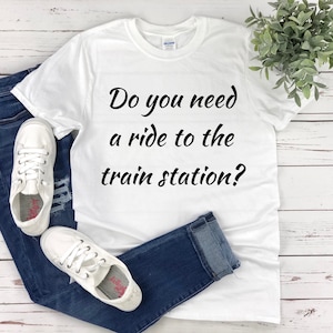 Könnte beinhalten: Weißes T-Shirt mit schwarzem Text, der lautet: "Brauchst du eine Mitfahrgelegenheit zum Bahnhof?"