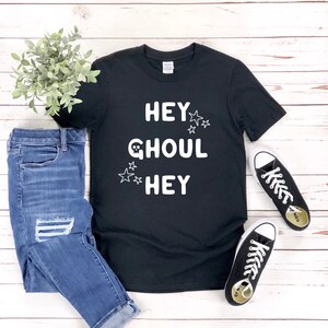 Peut inclure: T-shirt noir avec un texte blanc qui dit "Hey Ghoul Hey" avec un motif de crâne et des étoiles. Le t-shirt est associé à un jean bleu et des baskets blanches.