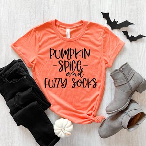 Peut inclure: Un t-shirt orange corail avec le texte "Pumpkin Spice and Fuzzy Socks" en lettres noires. Le t-shirt est posé sur une surface en bois blanc avec un jean noir, une citrouille blanche et deux chauves-souris noires.