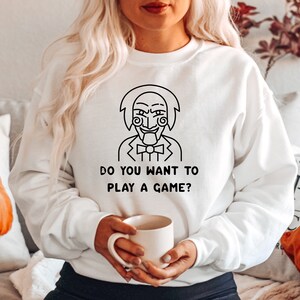 Peut inclure: Sweat-shirt blanc avec un dessin au trait noir d'un personnage du film Saw. Le texte "Do you want to play a game?" est imprimé sous le personnage.