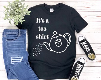 May include: Black t-shirt with white text that reads "It's a tea shirt" and a white outline of a teapot pouring tea.