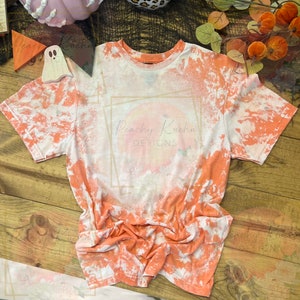 May include: A white and orange tie-dye t-shirt with a peach-colored circle and the text "Peachy Kuehn Designs" in a gold frame.