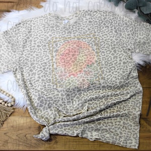 May include: A beige t-shirt with a leopard print pattern. The shirt has a graphic design of a peach with a gold frame.
