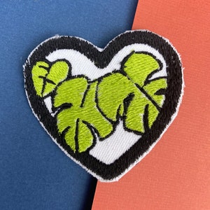 Könnte beinhalten: Ein grüner gestickter herzförmiger Patch mit schwarzem Rand und weißem Hintergrund. Der Patch zeigt ein Design aus zwei Monstera-Blättern.