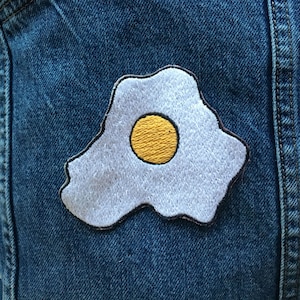 Puede incluir: Un parche blanco bordado con forma de huevo frito con una yema amarilla sobre un fondo de mezclilla azul.