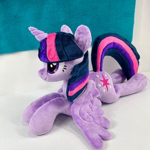 May include: A plush toy of Twilight Sparkle from My Little Pony. The toy is purple with a pink and purple mane and tail. It has a blue horn and a pink and purple rainbow symbol on its flank.