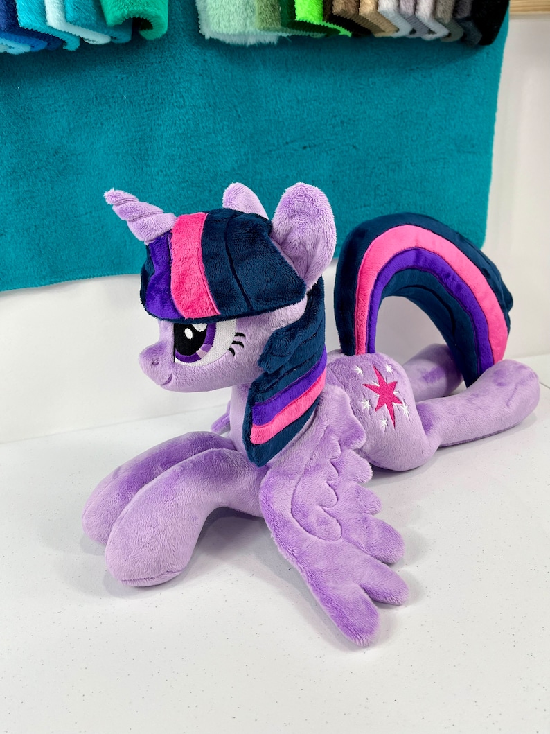 May include: A plush toy of Twilight Sparkle from My Little Pony. The toy is purple with a pink and purple mane and tail. It has a blue horn and a pink and purple rainbow symbol on its flank.