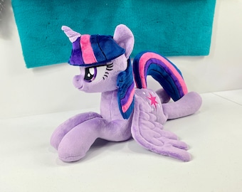 May include: A plush purple pony with a pink and blue mane and tail. The pony has a blue horn and a pink and blue star on its flank. The pony is lying on its side with its head turned to the left.