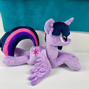 May include: A purple plush toy of Twilight Sparkle from My Little Pony. The toy is lying on its side with its wings spread out. It has a rainbow tail and a pink star on its flank.