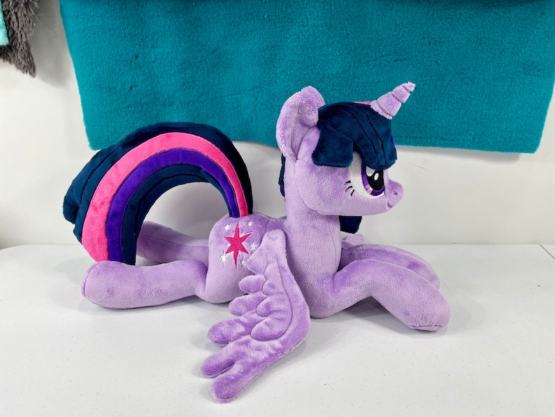 May include: A purple plush toy of Twilight Sparkle from My Little Pony. The toy is lying on its side with its wings spread out. It has a rainbow tail and a pink star on its flank.