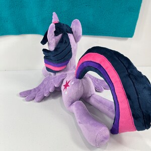 May include: Purple plush toy of Twilight Sparkle from My Little Pony. The toy is lying on its side with its wings spread out. The toy has a pink and purple tail and a dark blue mane.