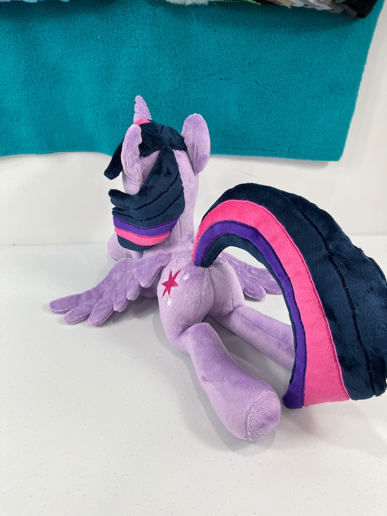 May include: Purple plush toy of Twilight Sparkle from My Little Pony. The toy is lying on its side with its wings spread out. The toy has a pink and purple tail and a dark blue mane.