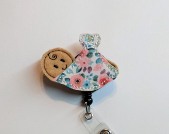 May include: A brown felt baby with a smiling face, wrapped in a white fabric with pink and blue floral print, attached to a black and silver retractable badge holder.