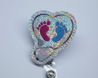 May include: A silver glitter heart-shaped badge holder with a blue and pink baby footprint design. The badge holder has a clear plastic strap and a silver swivel clip.
