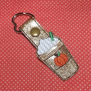 May include: Gold glitter keychain with a brown and white coffee cup with whipped cream and a pumpkin on top. The keychain has a heart-shaped ring.