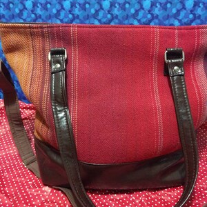 May include: A red and orange plaid tote bag with brown leather handles and a brown leather bottom. The bag has a long brown strap.