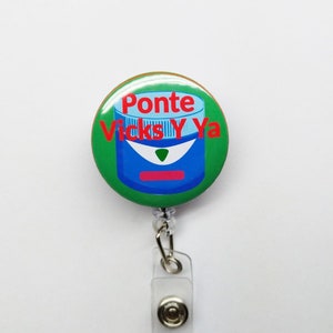 May include: A green and white badge reel with the text "Ponte Vicks Y Ya" in red. The badge reel features a blue illustration of a product container. The reel has a clear plastic strap and a metal clip.