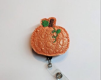 May include: A peach-colored pumpkin with green leaves and a brown stem, attached to a clear plastic retractable badge holder.