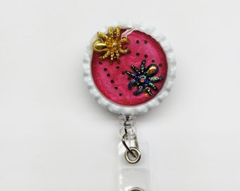 May include: A pink and white round badge holder with a black and gold spider and a blue and iridescent spider. The badge holder has a clear plastic retractable clip.