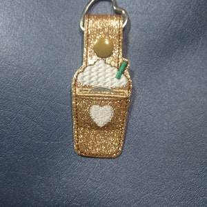 May include: A gold glitter keychain shaped like a coffee drink with a white heart and whipped cream design. The keychain has a silver heart-shaped ring and a green straw. The background is a dark blue surface.