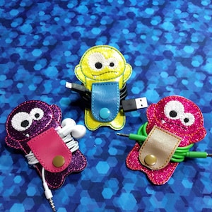 May include: Three colorful monster-shaped cord organizers. The first is purple with pink trim and white eyes, the second is yellow with blue trim and black eyes, and the third is pink with gold trim and white eyes. Each organizer has a button closure and is holding a cord.