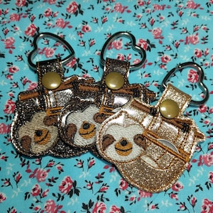 May include: Three keychains featuring a sloth design. The keychains are made of brown and gold fabric with glitter accents. The sloth faces are embroidered in white thread. Each keychain has a heart-shaped silver ring.